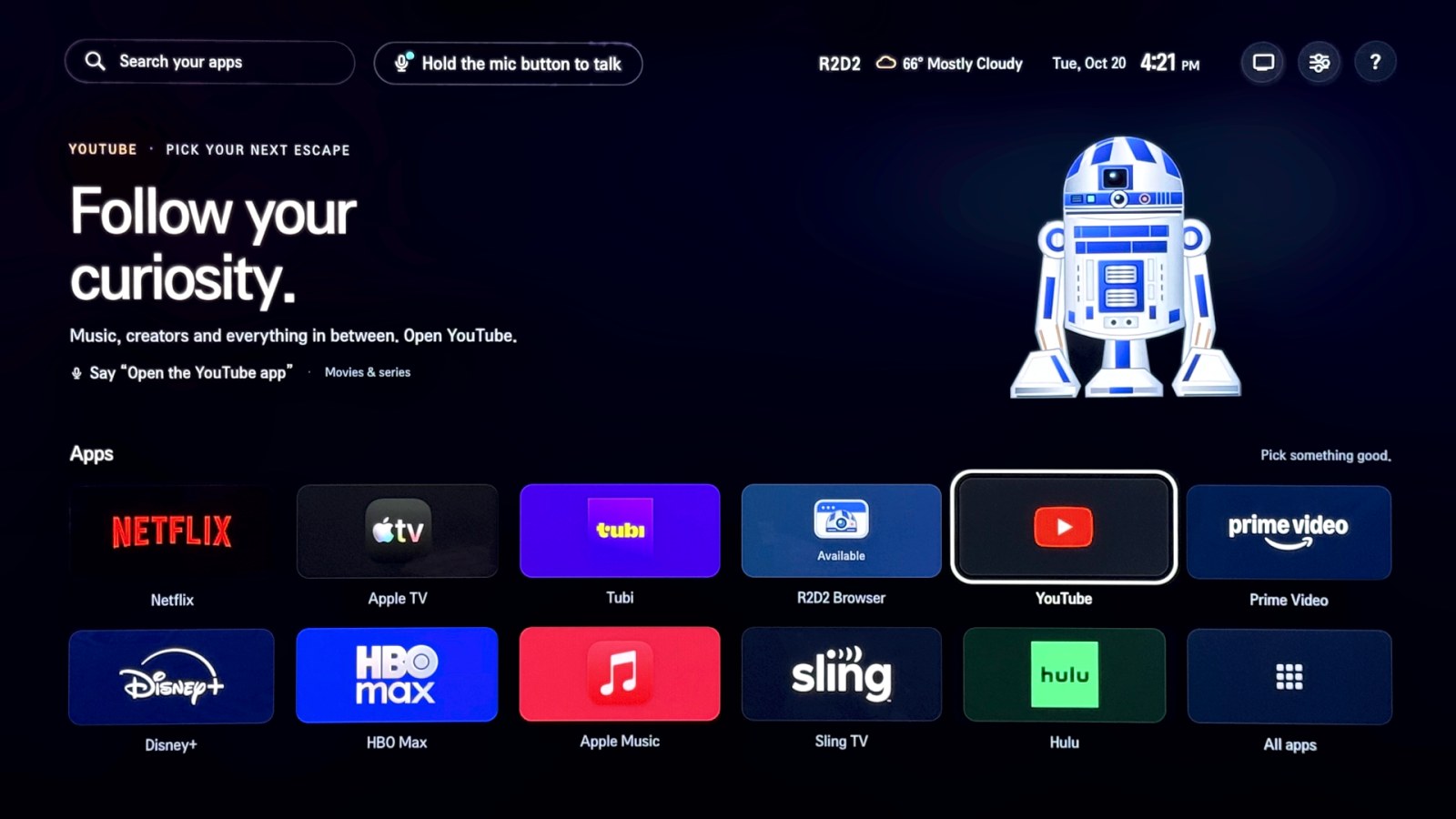 R2D2 Home on the TV: a search box and a 'Hold the mic button to talk' chip along the top, a hero reading 'Follow your curiosity.' for the focused YouTube tile beside the droid artwork, and a grid of twelve app tiles below.